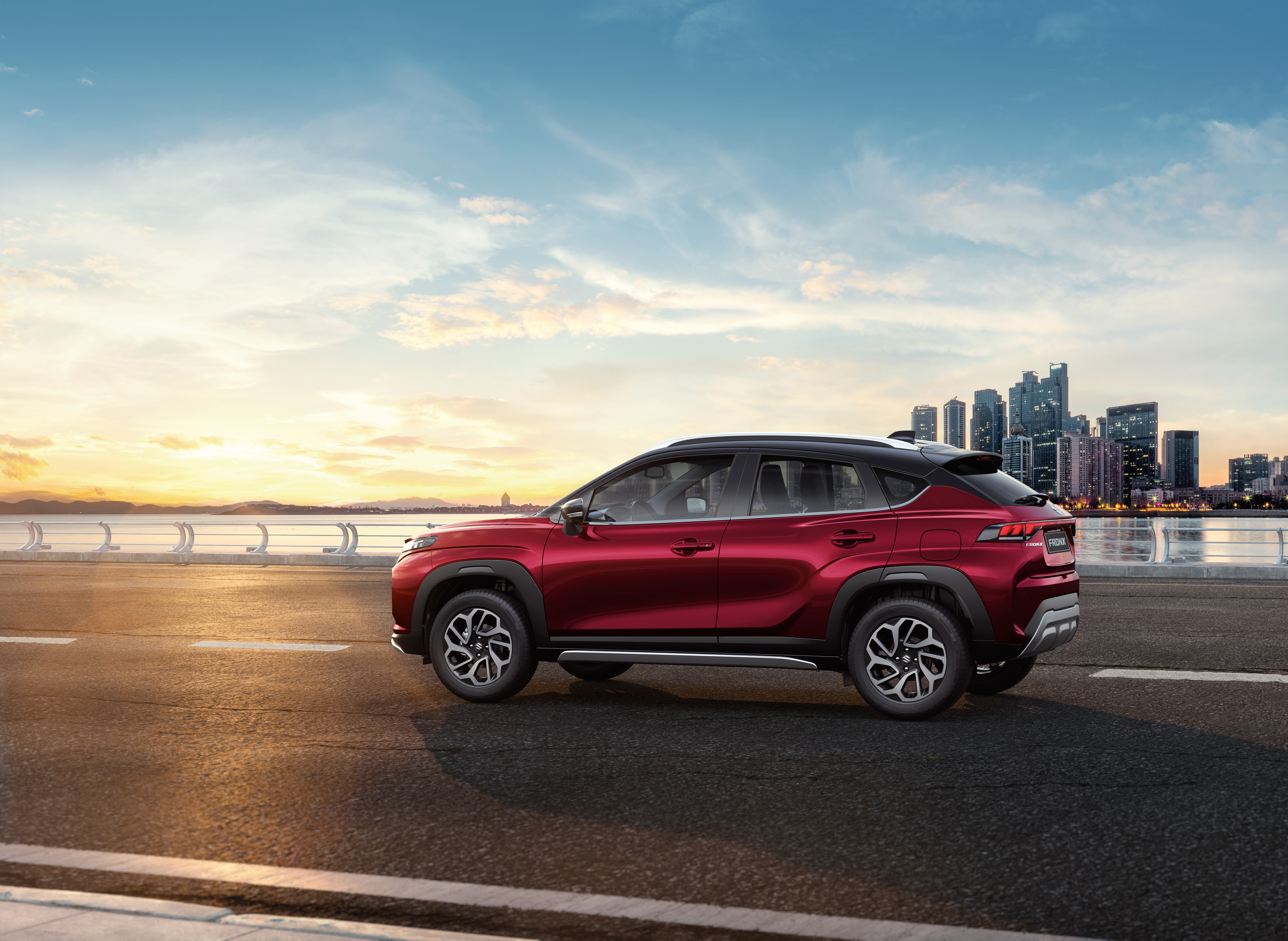 A red Suzuki Fronx compact SUV with a black roof parked on a waterfront road at sunset with a city skyline behind it