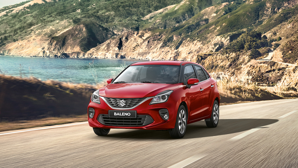 Suzuki Baleno driving by body of water with mountains in background